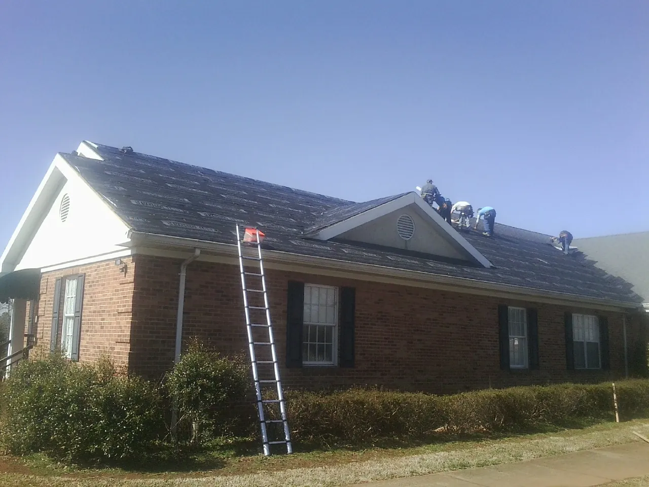 Crew tearing shingles off a brick ranch roof with a ladder set — Emergency Roof Repair work by our crew in Lakeland